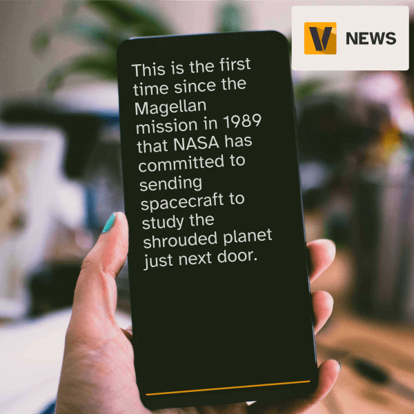 Blurred photo of a work table with a hand holding a smartphone in focus. The text on the screen reads, "This is the first time since the Magellan mission in 1989 that NASA has committed to sending spacecraft to study the shrouded planet just next door."