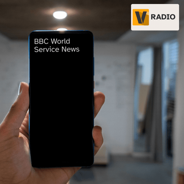 Image of a blurred room with a hand holding a smartphone in focus. The phone has a dark coloured screen. The text on the phone reads, "BBC World Service News".
