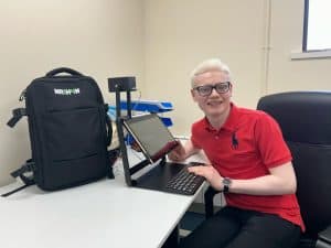 Nathan sits at a desk with his hand on the keyboard of the Nathan 14. You can see that the device has a 14 inch tablet with a distance camera attached. 